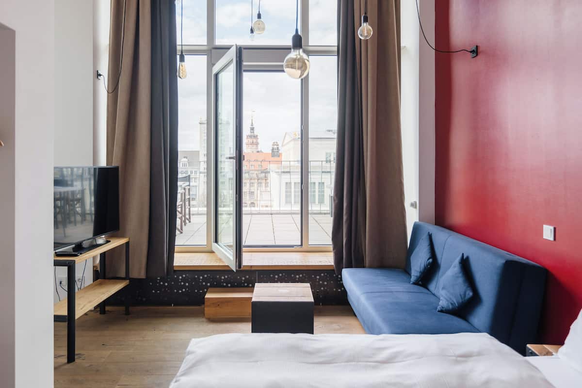 A modern hotel room with a large window, a sofa, a bed, a TV, and a wooden bench.