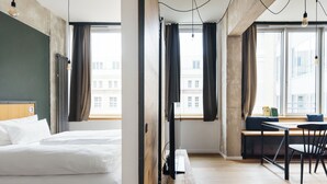 Une chambre d’hôtel moderne, dotée d’un lit, d’un bureau et d’une chaise, de grandes fenêtres avec des rideaux et de luminaires suspendus.