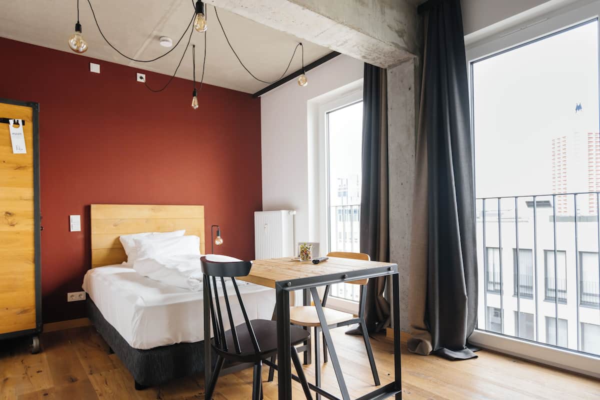 A hotel room with a bed, a wooden table, a chair, a radiator, and a window with curtains.
