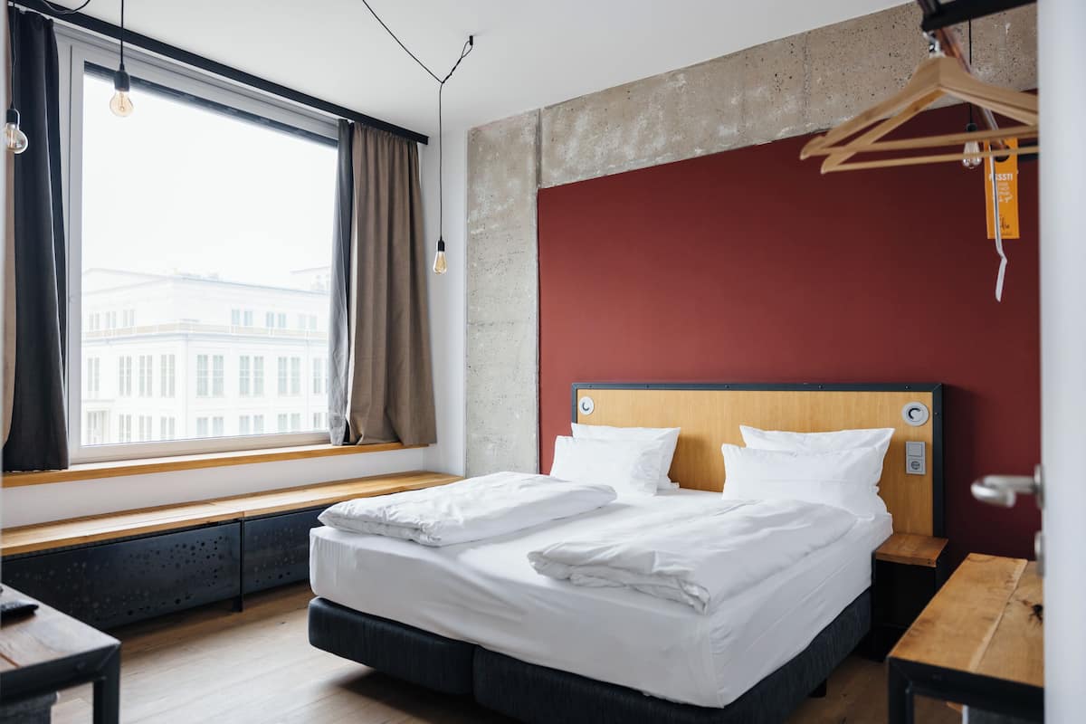 Habitación de hotel con una cama grande, un banco de madera, una mesa de madera, una lámpara colgante y vistas de edificios a través de una ventana.