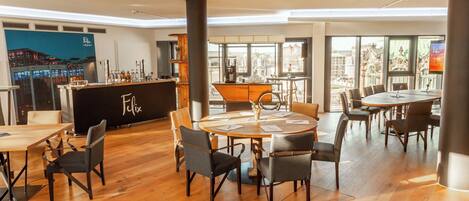 A modern hotel lobby with a bar area, round tables, and chairs, and large windows offering city views.