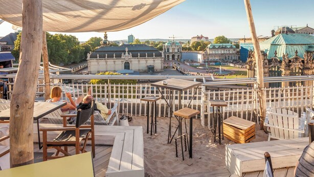 Eine Dachterrasse mit Holztischen und -stühlen, einem weißen Zaun und Blick auf historische Gebäude.