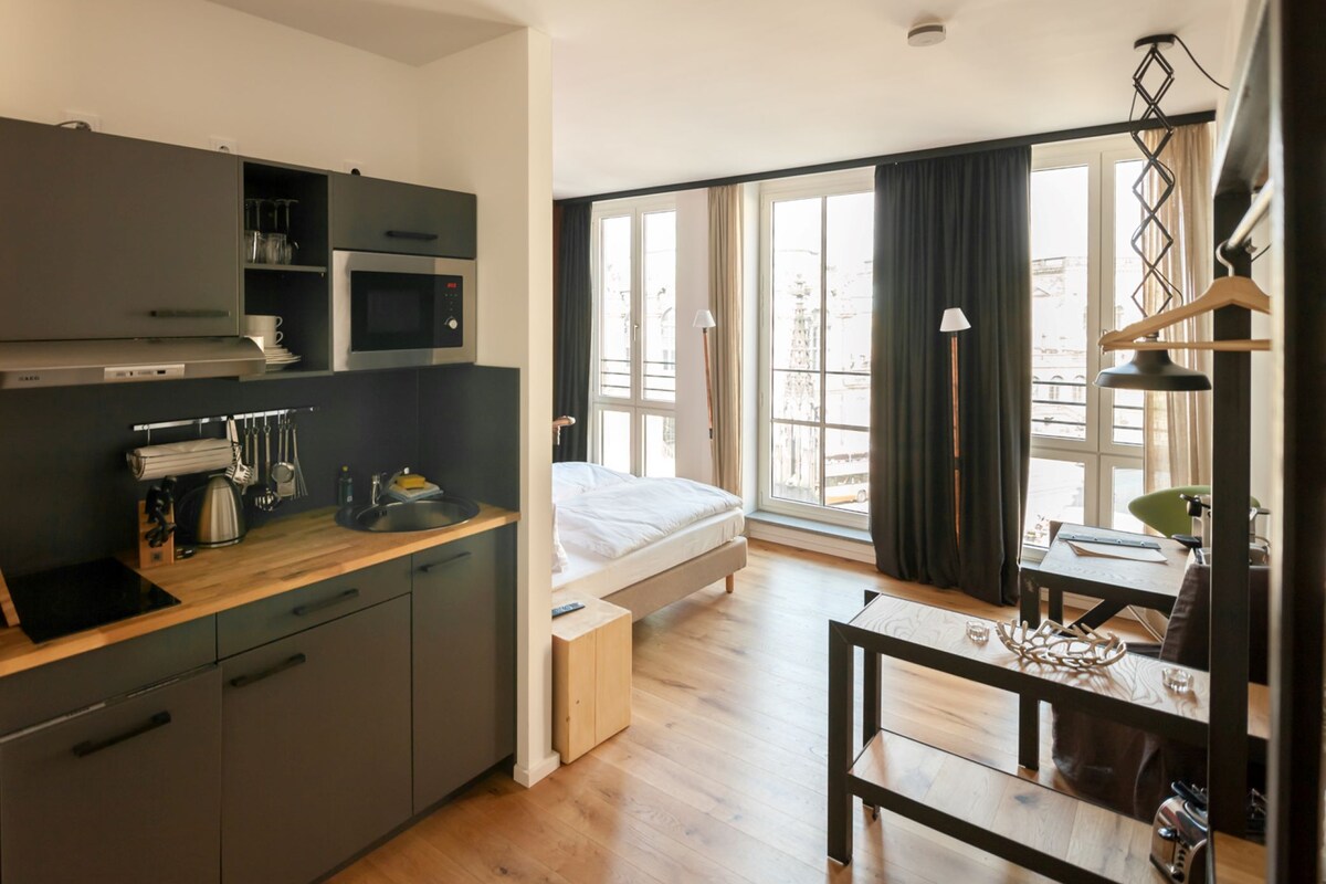 A compact hotel room with a kitchenette, a bed, a dining area, and large windows with curtains.