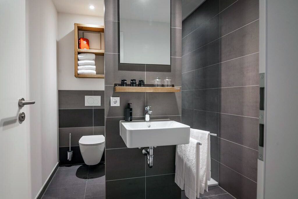 A modern bathroom with a white sink, dark tiled walls, a toilet, and a shelf with toiletries.