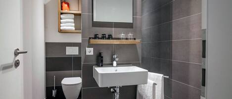A modern bathroom with a white sink, dark tiled walls, a toilet, and a shelf with toiletries.