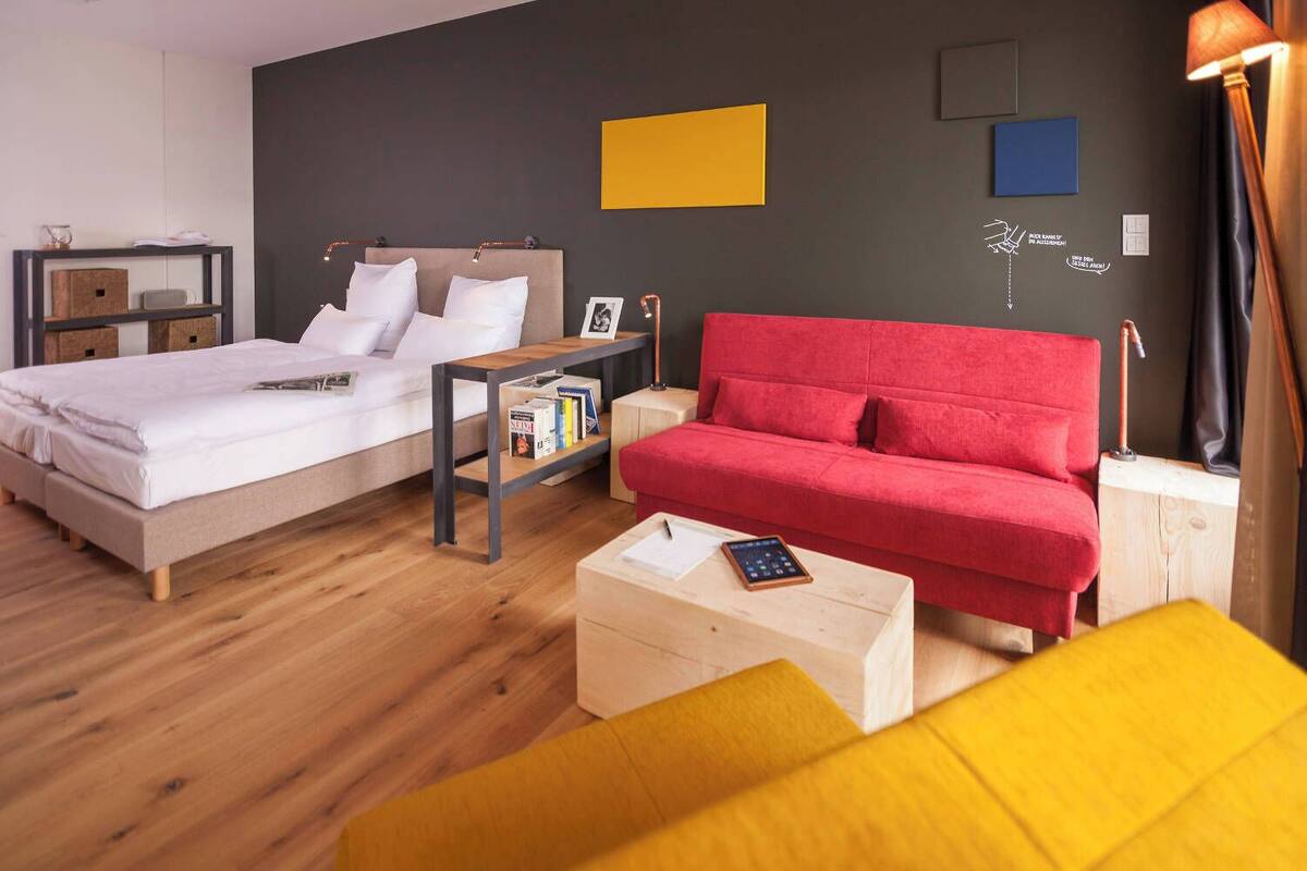 A modern hotel room with a bed, a red sofa, a wooden coffee table, and a nightstand with a lamp.