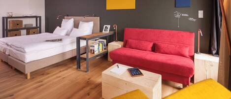 A modern hotel room with a bed, a red sofa, a wooden coffee table, and a nightstand with a lamp.