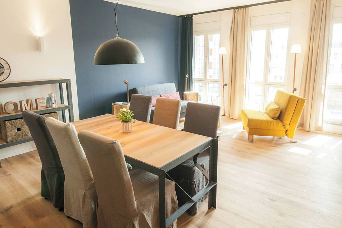 A modern living room with a wooden dining table, comfortable seating, a large window with curtains, and a dark blue accent wall.
