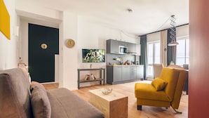 Ein modernes Wohnzimmer mit einer Couch, einem gelben Sessel und einem Couchtisch. Es gibt eine Küchenzeile mit einer Mikrowelle und Schränken.