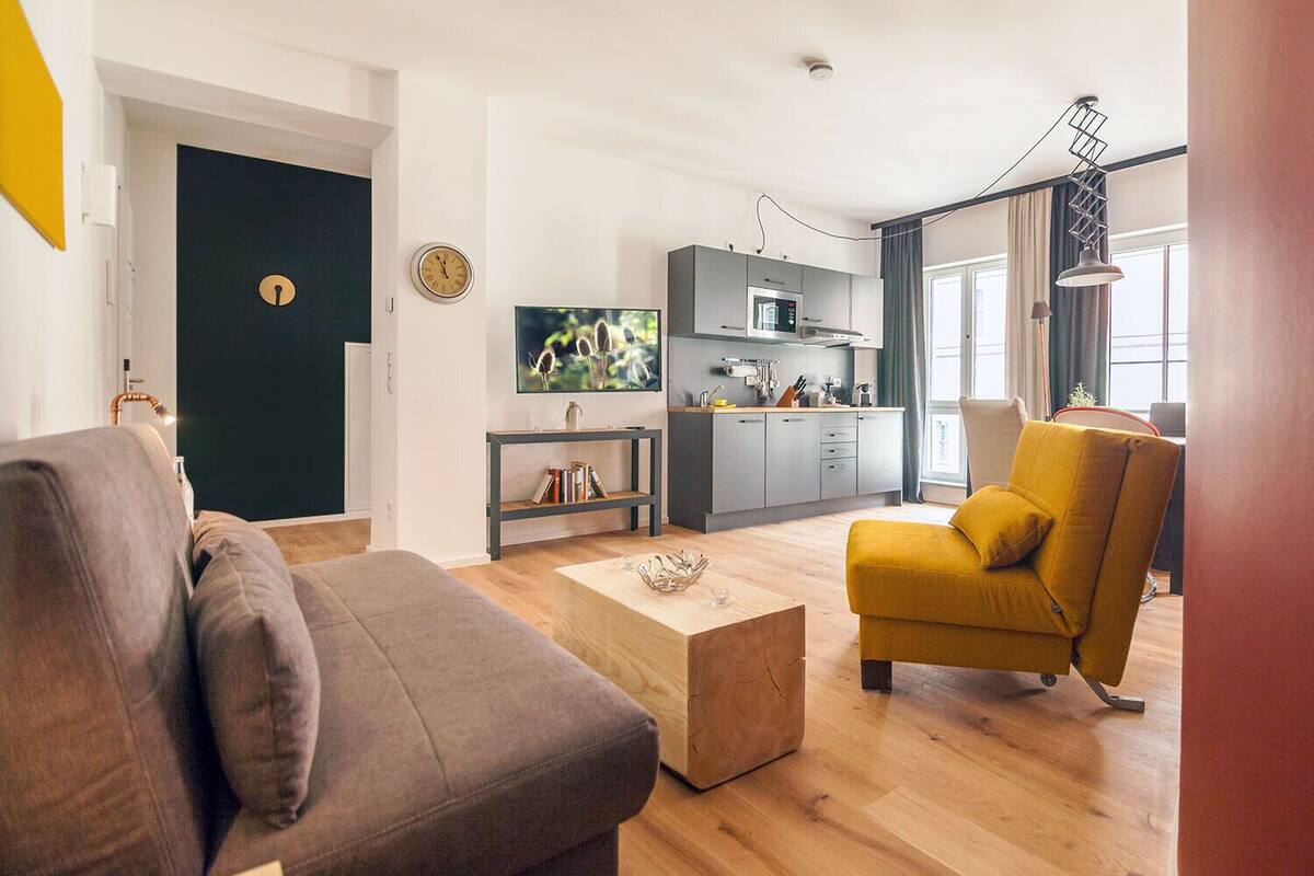 A modern living room with a sofa, a yellow armchair, and a coffee table. There is a kitchen area with a microwave and cabinets.