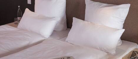 Una cama bien hecha con sábanas y almohadas blancas, una botella de agua y una revista en la mesita de noche.