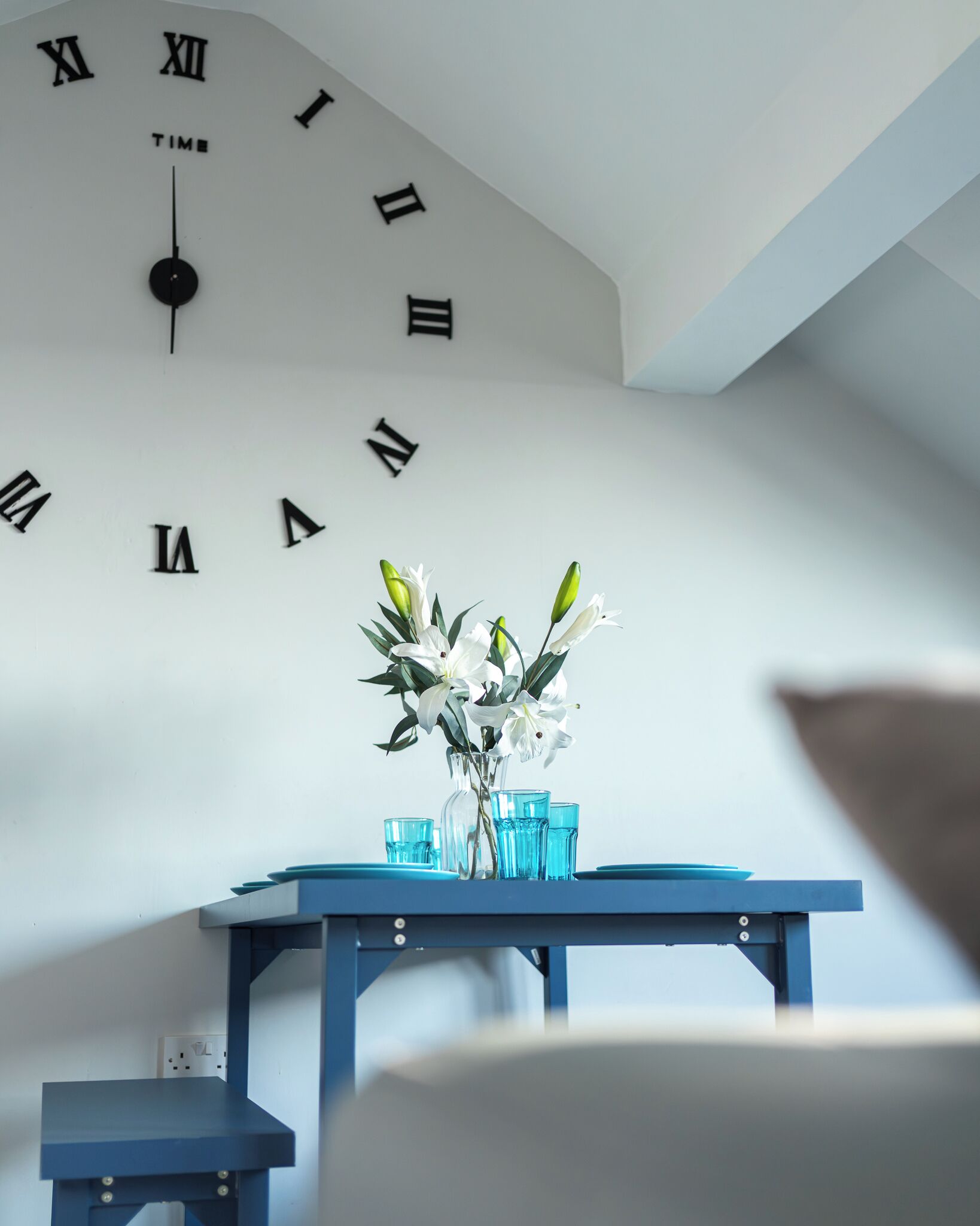 Una habitación con un reloj de pared mostrando números romanos, un florero con flores y una mesa azul con vasos.
