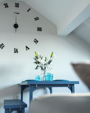 Une pièce avec une horloge murale affichant des chiffres romains, un vase rempli de fleurs et une table bleue sur laquelle sont posés des verres.