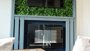 A modern living room with a flat-screen TV mounted above a fireplace, surrounded by green foliage. - Vibe Apartment 4, Lord Street (Blackpool)