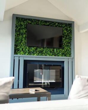 A modern living room with a flat-screen TV mounted above a fireplace, surrounded by green foliage.