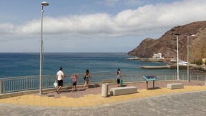 Un paseo marítimo con barandilla, bancos y una vista despejada del mar y una colina rocosa al fondo.