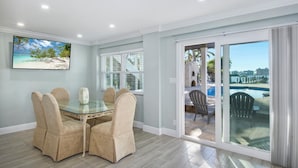 A dining area with a glass table and beige chairs, a large window with blinds, and a view of a pool and outdoor seating.