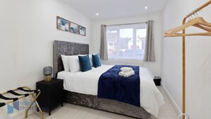 A bedroom with a bed, a bedside table, a hanging coat rack, and a window with curtains. - Comfy 4 Bedroom house, Ideal for Workers and Families, FREE Parking (Coulsdon)