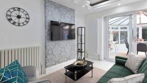 A modern living room with a green sofa, a black coffee table, a wall-mounted TV, and a decorative clock. - Comfy 4 Bedroom house, Ideal for Workers and Families, FREE Parking (Coulsdon)