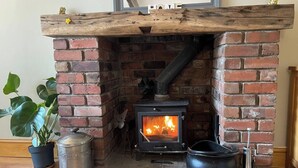A brick fireplace with a wood-burning stove, a potted plant, and a metal pot on the floor. - 2bed Cottage - Trawden - Pet Friendly - Sleeps4 (Colne)