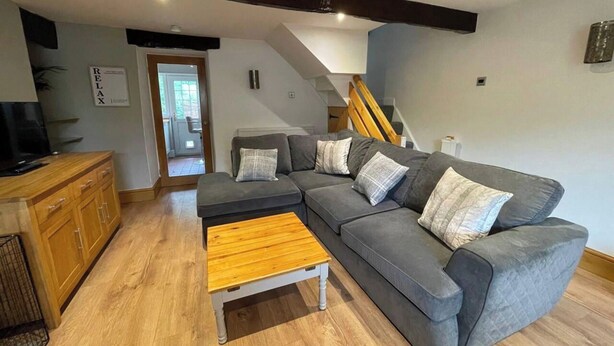 A modern living room with a grey sectional sofa, a wooden coffee table, and a wooden cabinet. There is a staircase leading to an upper level. - 2bed Cottage - Trawden - Pet Friendly - Sleeps4 (Colne)
