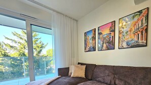 A living room with a large window, a brown sofa, and three framed paintings on the wall. - Apartament Buena Vista - 365PAM (Kolobrzeg)