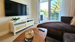 A modern living room with a flat-screen TV, a sofa, a coffee table with a glass vase, and a view of trees and buildings through large windows. - Apartament Buena Vista - 365PAM (Kolobrzeg)