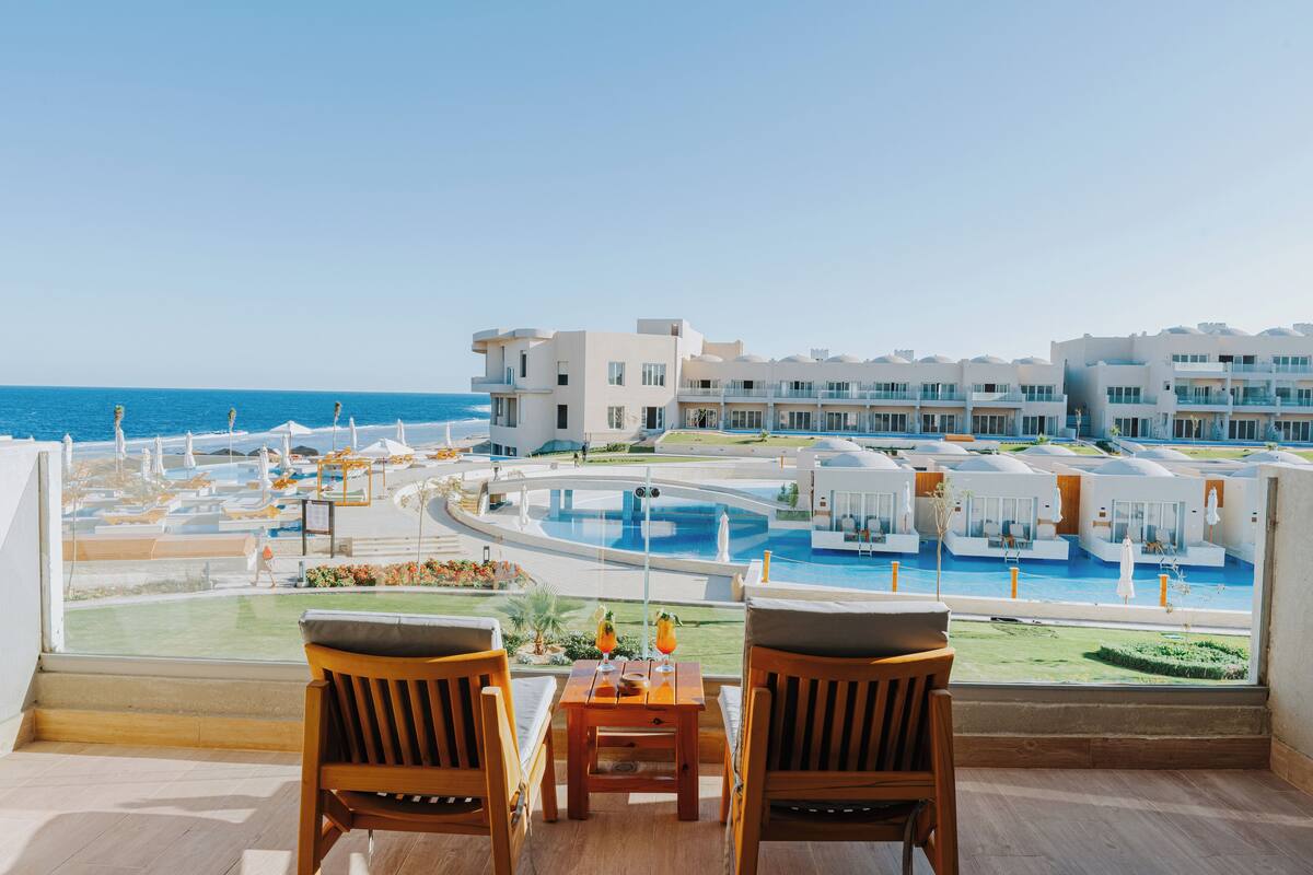 Ein Balkon mit Holzsesseln und einem Tisch, mit Blick auf einen Poolbereich mit mehreren Gebäuden und freiem Meerblick.