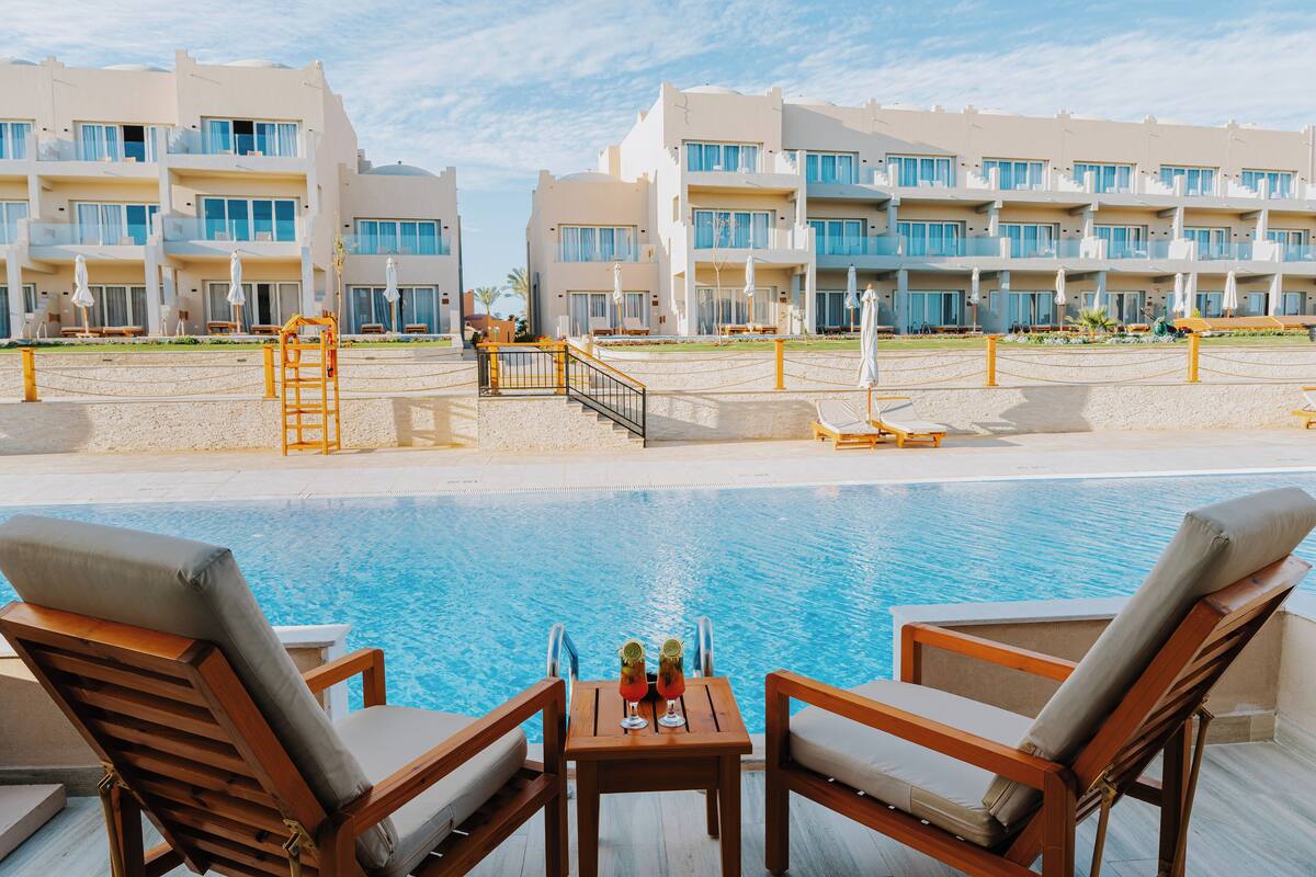 Ein Poolbereich mit Liegestühlen und einem Tisch, ein klares, blaues Schwimmbecken und ein Hotelgebäude im Hintergrund.