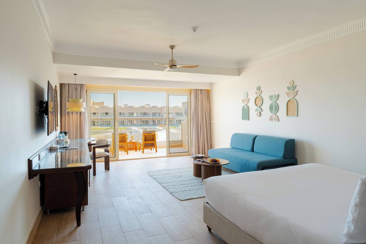 Ein Hotelzimmer mit einem Bett, einem Schreibtisch, einem blauen Sofa, einem Essbereich mit Stühlen und Blick auf Gebäude durch die Glastüren.