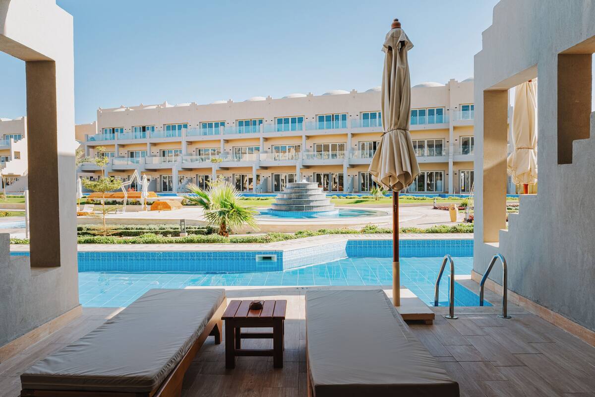 Ein Poolbereich mit Liegestühlen, einem kleinen Tisch und Blick auf ein Hotelgebäude im Hintergrund.