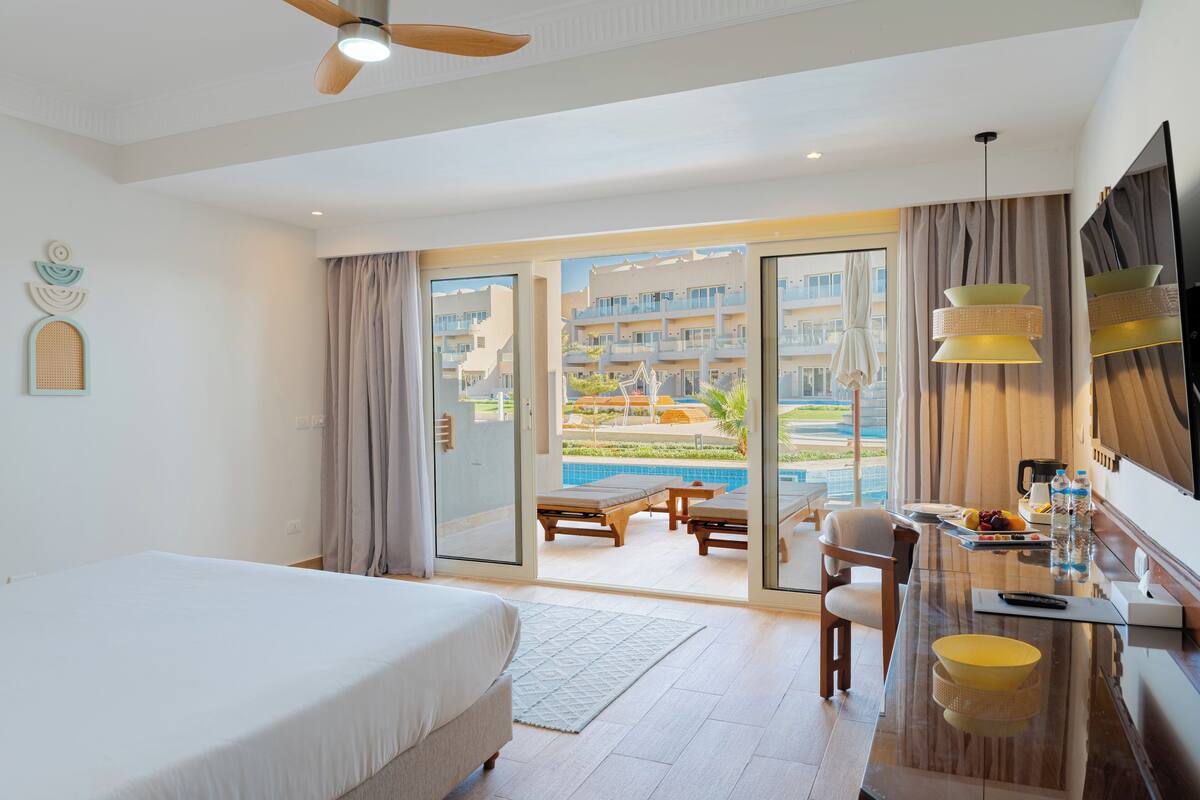 Ein Hotelzimmer mit Bett, Schreibtisch mit Stuhl, Fernseher und Blick auf einen Pool sowie Gebäude.
