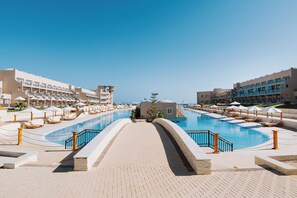 Zona de piscina en un resort, con vista clara a la piscina, sillones de descanso y un edificio con varios balcones.