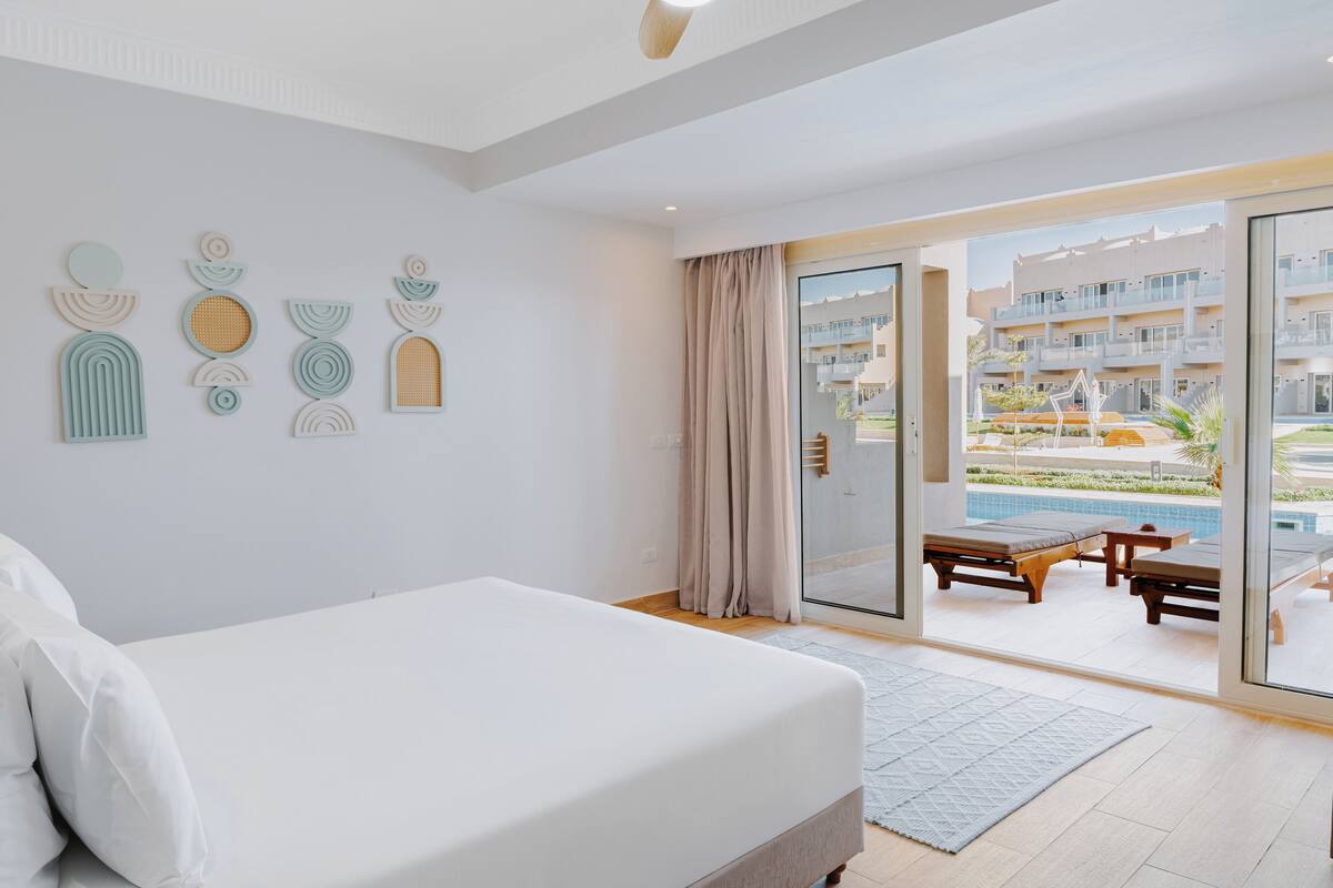 Ein modernes Hotelzimmer mit einem großen Bett, Blick auf den Pool und Sitzgelegenheiten im Freien sowie geometrischen Wandbildern.