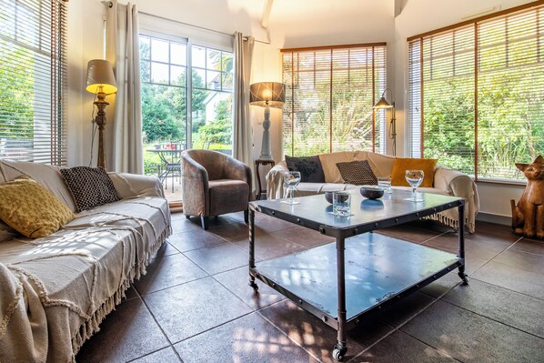 A cozy living room with a sofa, armchair, and coffee table, large windows with blinds, and a view of greenery outside.