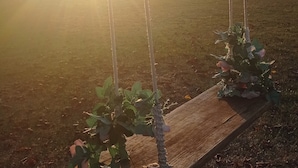 A wooden swing set with ropes, positioned on a grassy field with a sunset in the background.