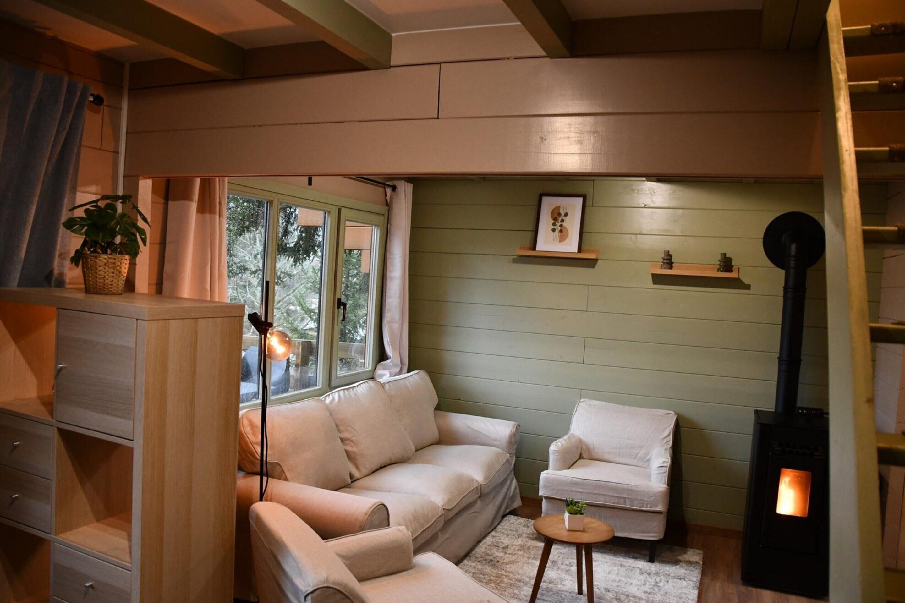A cozy living room with a beige sofa, a white armchair, a small table with a plant, a fireplace, and a window with curtains.
