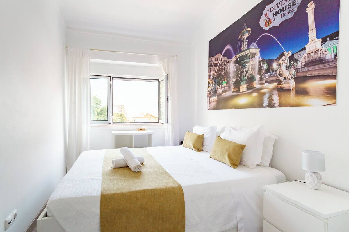 Una habitación de hotel con una cama grande, una mesita de noche, una lámpara, una ventana con cortinas y un cartel con una vista urbana.