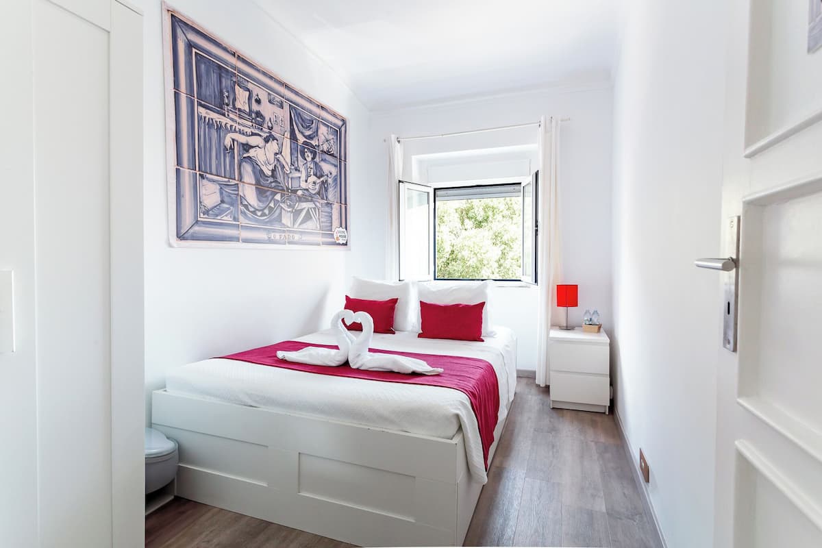 Un dormitorio con cama, mesita de noche, ventana y un cuadro colgado en la pared.
