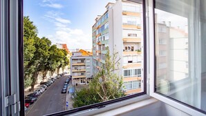 A view from a window of a multi-story building with balconies, parked cars on the street, and trees.
