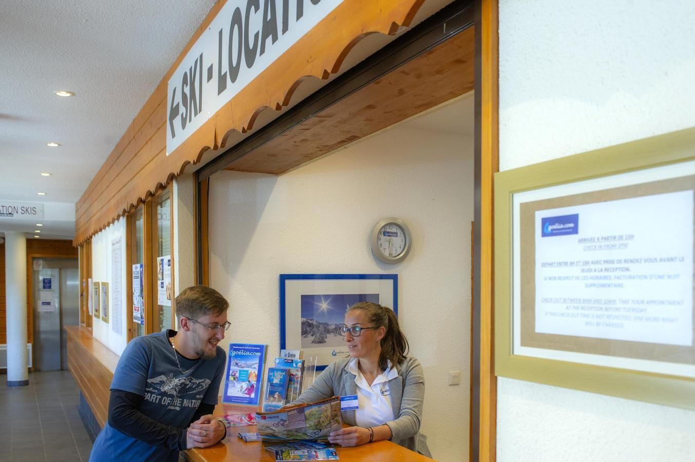 Un comptoir d’accueil avec une pancarte au-dessus indiquant « LOCATION DE MATÉRIEL DE SKI ». Des affiches d’information sont accrochées au mur, et il y a une horloge.