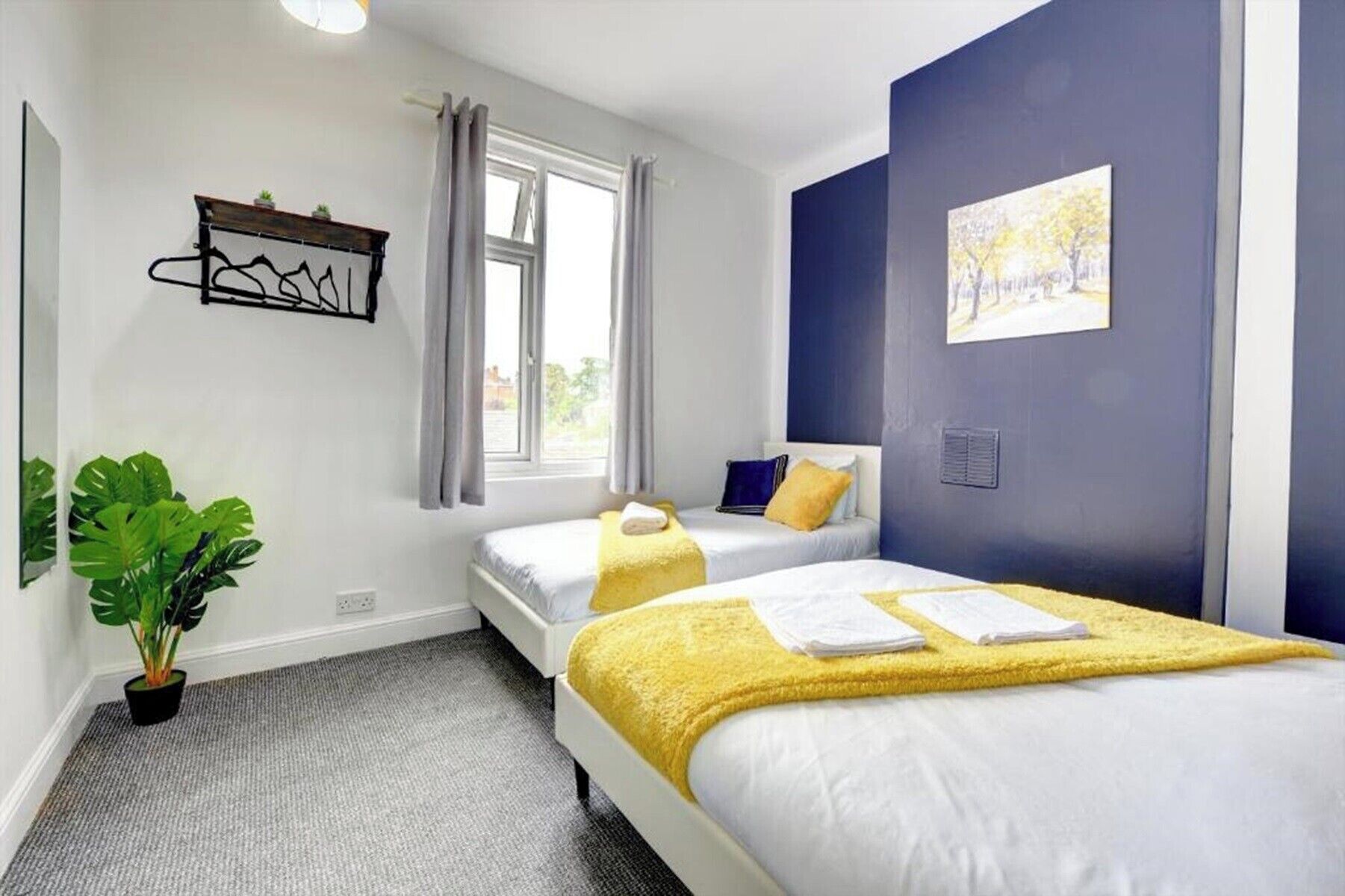 Hotellihuone, jossa on kaksi sänkyä, ikkuna, jossa on verhot, huonekasvi ja seinälle kiinnitetty hylly.