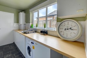 A modern kitchen with a built-in washing machine, a window with a view of the outside, and a wall clock. - Central - 2br House - Free Parking - Garden (Coventry)