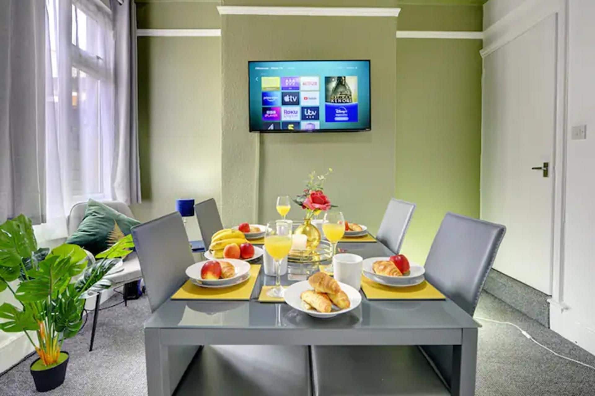 Ein Essbereich mit einem Tisch für zwei Personen, einem Laptop und einem an der Wand montierten Flachbildfernseher, der verschiedene Sender zeigt.