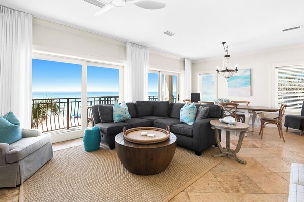 A spacious living room with a large window overlooking the sea, a dark-colored sectional sofa, a round coffee table, and a dining area with wooden furniture.