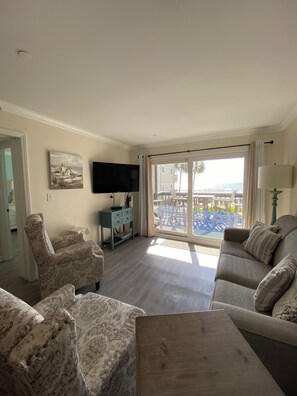A living room with a sofa, armchair, and a flat-screen TV. There is a large window with a view of the outdoors.