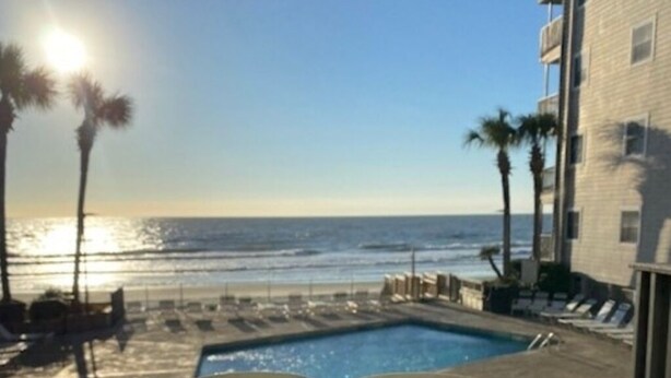 A poolside view with a clear view of the ocean, palm trees, and a building with a balcony.
