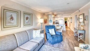 A spacious living room with a grey sofa, a blue armchair, and a coffee table. There are framed artworks on the walls and a chandelier hanging from the ceiling.