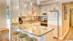 A kitchen with granite countertops, white cabinets, a refrigerator, and a sink. There are three bar stools with green cushions.