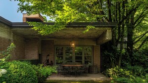 A covered patio with a table and chairs, surrounded by lush greenery and a brick building. - Wisteria Holiday Home (Lonato)
